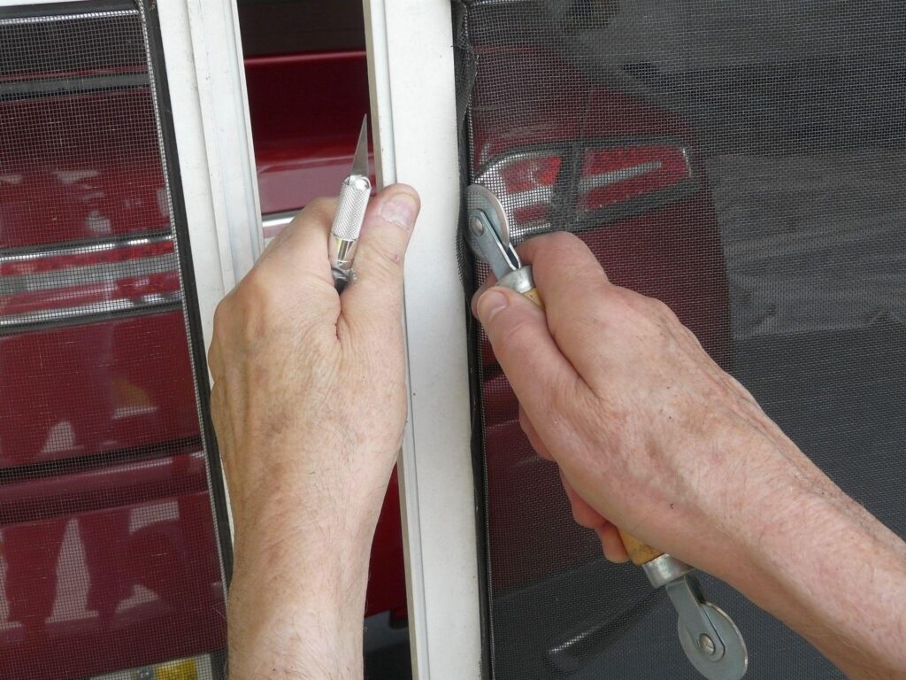 A homeowner is using a spline roller tool and knife to replace a damaged panel in a sliding screen door